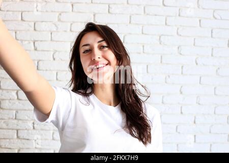 Indoor portrait of stunning beautiful girl making selfie on mobile cell phone, trendy urban 90s hipster style, funny positive emotions, long hair & nu Stock Photo
