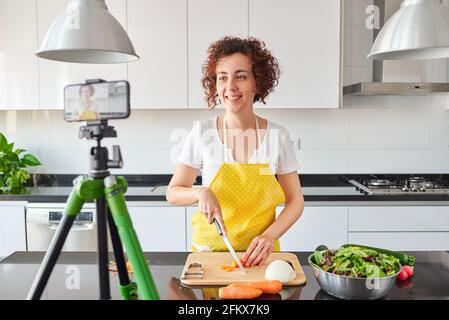 Woman youtuber records herself with her smartphone in her kitchen while preparing a salad recipe, there is natural light and she is wearing a yellow a Stock Photo