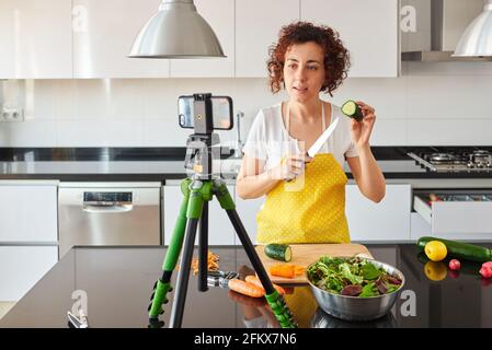 Woman youtuber records herself with her smartphone in her kitchen while preparing a salad recipe, there is natural light and she is wearing a yellow a Stock Photo