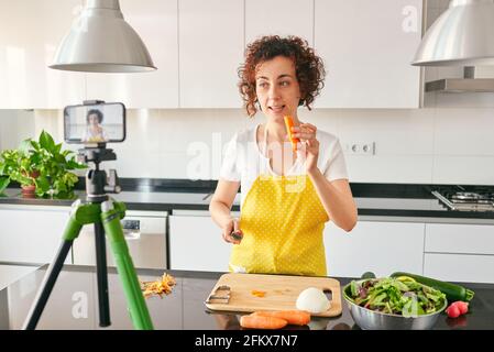 Woman youtuber records herself with her smartphone in her kitchen while preparing a salad recipe, there is natural light and she is wearing a yellow a Stock Photo