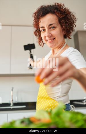 Woman youtuber records herself with her smartphone in her kitchen while preparing a salad recipe, there is natural light and she is wearing a yellow a Stock Photo