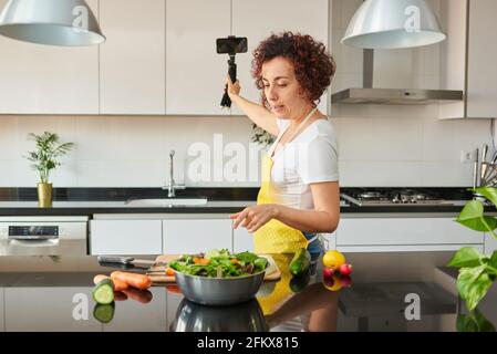 Woman youtuber records herself with her smartphone in her kitchen while preparing a salad recipe, there is natural light and she is wearing a yellow a Stock Photo
