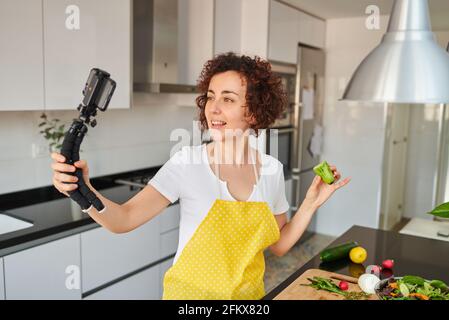 Woman youtuber records herself with her smartphone in her kitchen while preparing a salad recipe, there is natural light and she is wearing a yellow a Stock Photo