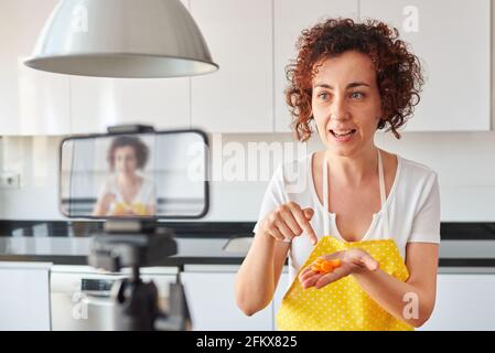 Woman youtuber records herself with her smartphone in her kitchen while preparing a salad recipe, there is natural light and she is wearing a yellow a Stock Photo