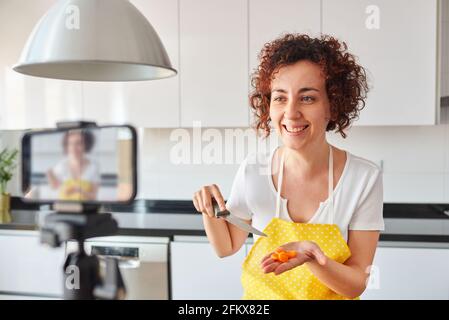 Woman youtuber records herself with her smartphone in her kitchen while preparing a salad recipe, there is natural light and she is wearing a yellow a Stock Photo