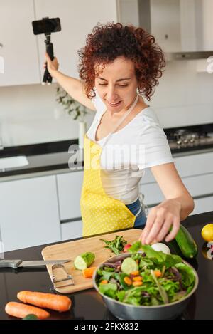 Woman youtuber records herself with her smartphone in her kitchen while preparing a salad recipe, there is natural light and she is wearing a yellow a Stock Photo