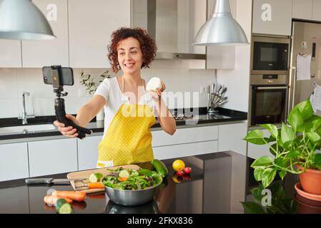 Woman youtuber records herself with her smartphone in her kitchen while preparing a salad recipe, there is natural light and she is wearing a yellow a Stock Photo