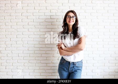 Young attractive woman wearing white shirt & cat eye glasses smiling wide. Pretty female model, hands crossed on chest, brunette hair posing. Girl, wh Stock Photo