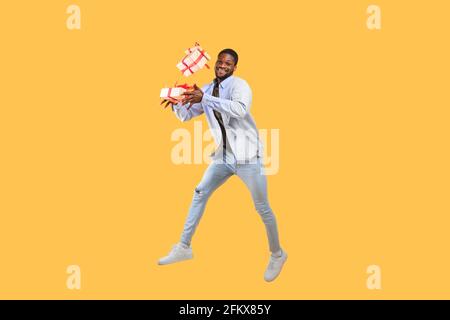 Holidays and celebration time. Overjoyed african american guy holding gift boxes and jumping on yellow background Stock Photo