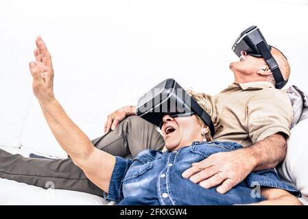 Senior mature couple having fun together with virtual reality headset sitting on sofa - Happy retired people using modern vr goggle glasses - New tren Stock Photo