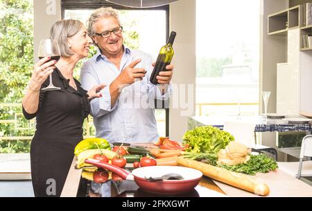 Senior couple cooking healthy food and drinking red wine at house kitchen - Retired people at home preparing lunch with fresh vegetables and bio produ Stock Photo