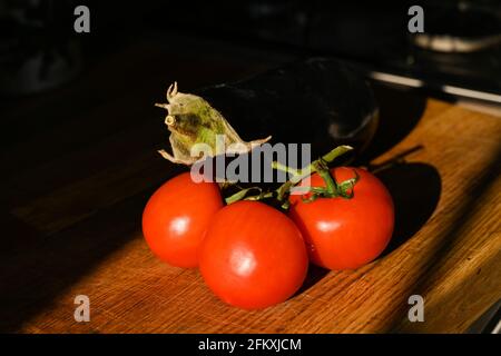 Tomatoes on the vine and a fresh aubergine (eggplant) Solanum melongena on a wooden chopping board Stock Photo