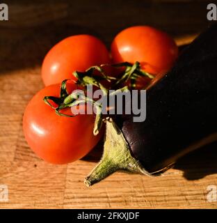 Tomatoes on the vine and a fresh aubergine (eggplant) Solanum melongena on a wooden chopping board Stock Photo