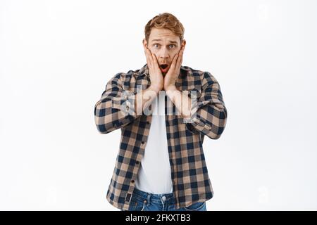 Shocked and concerned redhead man gasping, holding hands on face and staring speechless startled, nervous about accident, hear bad news, standing over Stock Photo