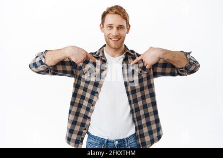 Smiling redhead man pointing at center and looking happy, showing logo on t-shirt, pointing at himself, standing against white background Stock Photo