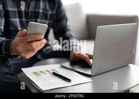 Unrecognizable Business Guy Using Phone And Laptop Indoors, Cropped Stock Photo