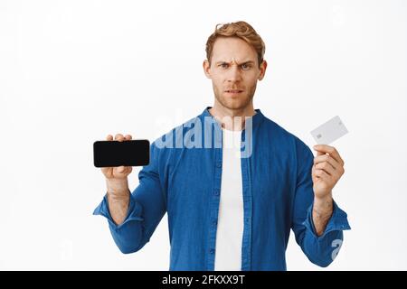 Disappointed young man shows smartphone screen flipped horizontal and bank credit card, frowning upset or angry, looking displeased as showing bad app Stock Photo