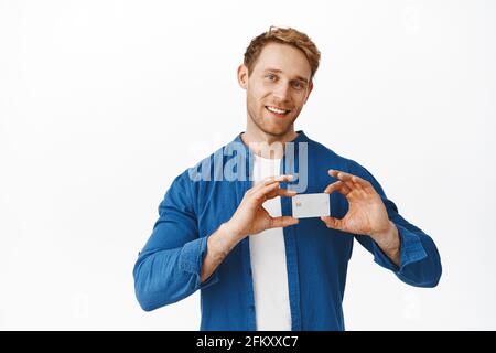 Image of handsome redhead man shows credit card near his heart, love and like bank, smiling pleased, recommending company, standing over white Stock Photo