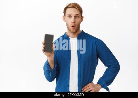 Surprised redhead man showing application on smartphone screen. Guy shows mobile phone display and say wow with amazed face, standing over white Stock Photo