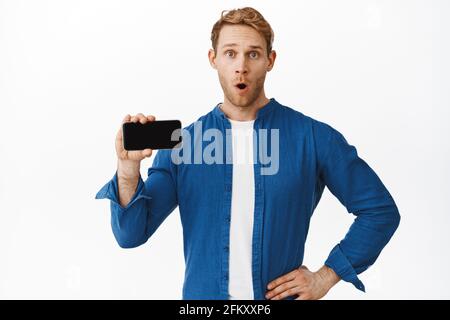 Guy looks surprised while shows smartphone empty screen, gasping and saying wow, staring amazed, found something interesting online, standing over Stock Photo
