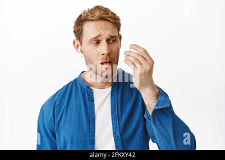 Man looks confused on wireless earphone, broken earbud while listening music, talking in headphones, standing against white background Stock Photo