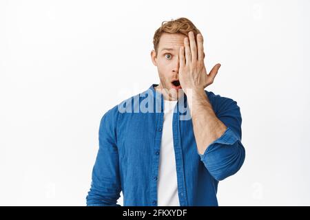 Close up of redhead male model looking with one eye side of face and drop jaw, gasping amazed, say wow, checking out awesome advertisement offer Stock Photo