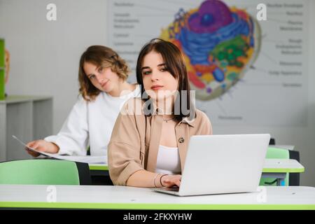 Two beautiful high school female teenage friends sitting on desks in modern class with laptop and notebook and look at camera Stock Photo