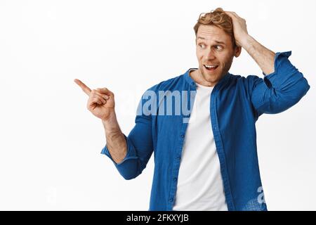 Surprised redhead man touch head, pointing and looking left at copyspace banner with startled face, standing against white background Stock Photo