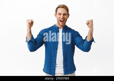 Encouraged and determined redhead man scream yeah, shaking clenched hands and shouting courageous, excited of winning prize, achieve goal, celebrating Stock Photo