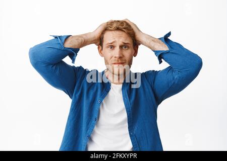Troubled and worried young redhead man, look distressed, holding hands on head indecisive and nervous, panicking, having problem, standing upset and Stock Photo