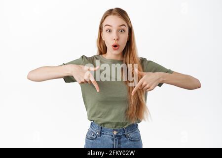 Image of intrigued blond girl gasp, say wow and points fingers down at interesting advertising promo offer, asks question about product, stands over Stock Photo