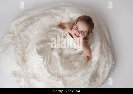 Toddler girl looks up and smiles while wearing mother's wedding gown