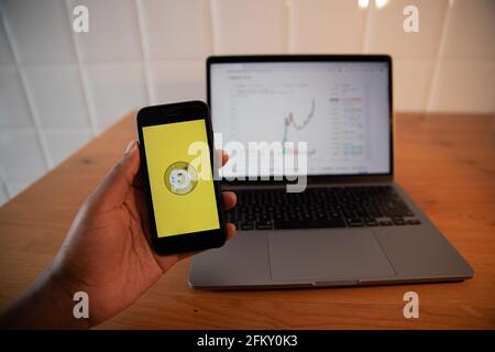 New York, May 4th 2021: Close up of the Dogecoin logo on the iPhone display standing in front of a macbook with a graphic of its value. Dogecoin is a Stock Photo