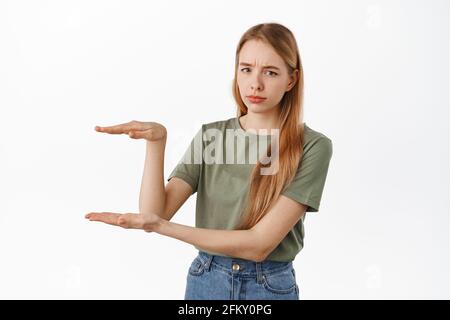 Image of skeptical blond girl, holding empty copyspace, looking unsure and doubtful, frowning displeased, display an item against white background Stock Photo