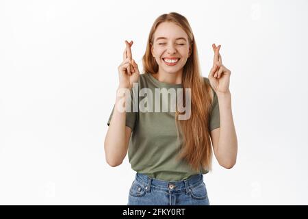 Image of happy optimistic blond girl, close eyes and cross fingers for good luck, praying, waiting for fortune good sign, begging for wish come true Stock Photo