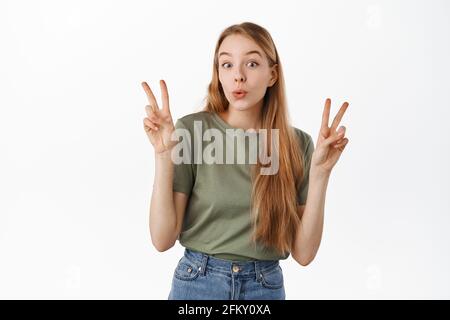 Cute and funny blond girl shows peace v-sign and pucker lips silly, standing happy in summer t-shirt and jeans against white background Stock Photo