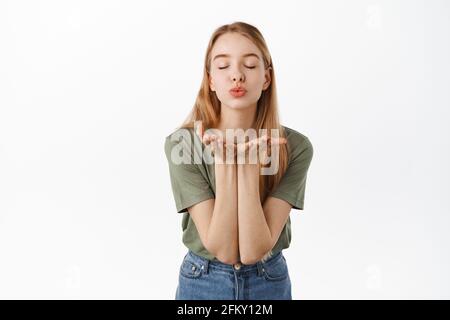 Romantic cute girlfriend sending air kiss at camera with closed eyes, puckered lips and hands near mouth, standing over white background Stock Photo