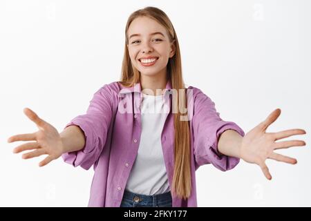 Image of beautiful blond girl reaching for something, stretch out hands to hold, hug or receive, smiling happy and friendly, standing over white Stock Photo