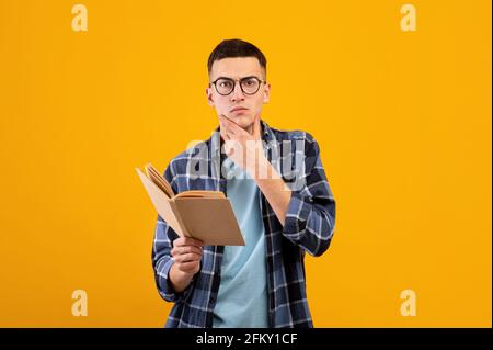 Pensive student in casual wear reading textbook, thinking over complicated task, studying for exam on orange background Stock Photo