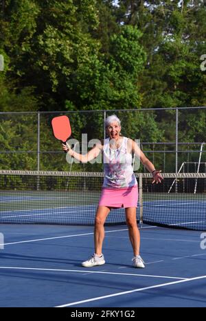 Female Pickle Ball player shows her love and excitement for the game.  She is standing on a tennis court holding a red racket. Stock Photo