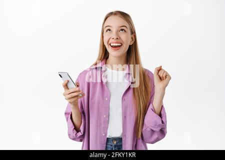 Thank god. Smiling happy girl looking up relieved after winning on mobile phone, holding smartphone and rejoicing, celebrating achievement in app Stock Photo