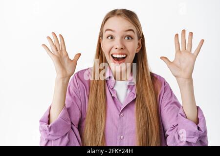 Happy surprised girl reacts to super cool news, raising hands up, smiling and gasping amazed, staring excited at camera, standing against white Stock Photo
