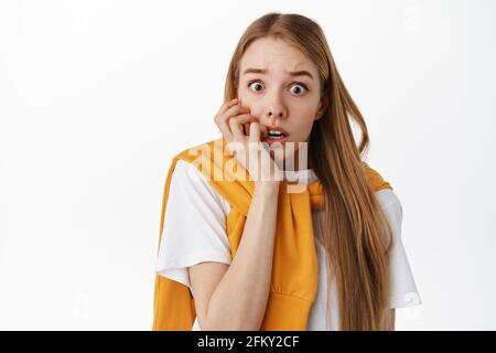 Close up of shocked and scared blond woman, biting fingers and gasping, staring startled and frightened, afraid of something scary, standing over Stock Photo