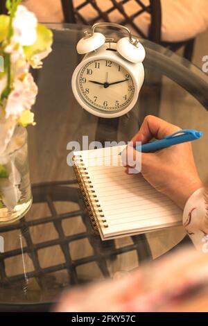 Personal diary concept with woman writes in notebook, cozy workspace glass table with plant and alarm clock, over shoulder view Stock Photo