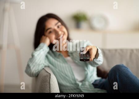 Happy Indian lady switching TV channels with remote control on sofa at home, selective focus Stock Photo