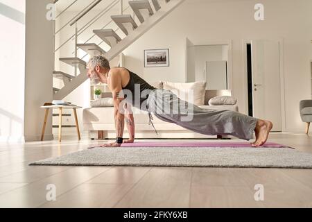 Fit tattooed mature man doing plank morning workout yoga exercise at home. Stock Photo