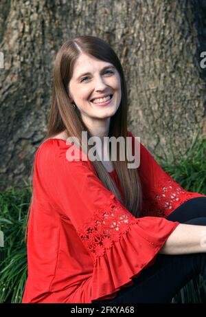 Happy smile lights this young woman's face.  She is sitting outsides on the grass besides a tree.  Her shirt is red and she has long brown hair. Stock Photo