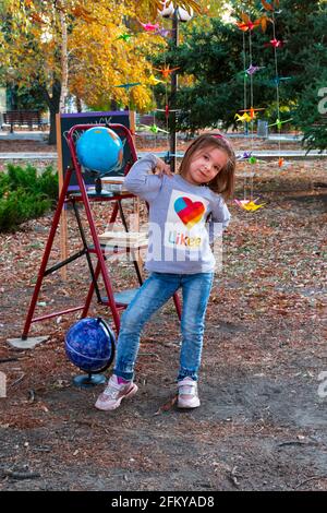 Adorable little schoolgirl against the background of an autumn park against the background of a school board and a globe. Back to school. Stock Photo