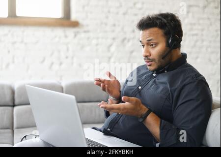 Concentrated indian man wearing headset holding virtual video conference sitting on the couch at home with a laptop computer on the laps, multiethnic guy studying online, involved webinar Stock Photo