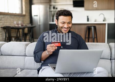 Happy indian guy holding credit card and paying online, using laptop computer for shopping online, transferring money, mixed-race man sits on the couch and input banking card data for purchasing Stock Photo